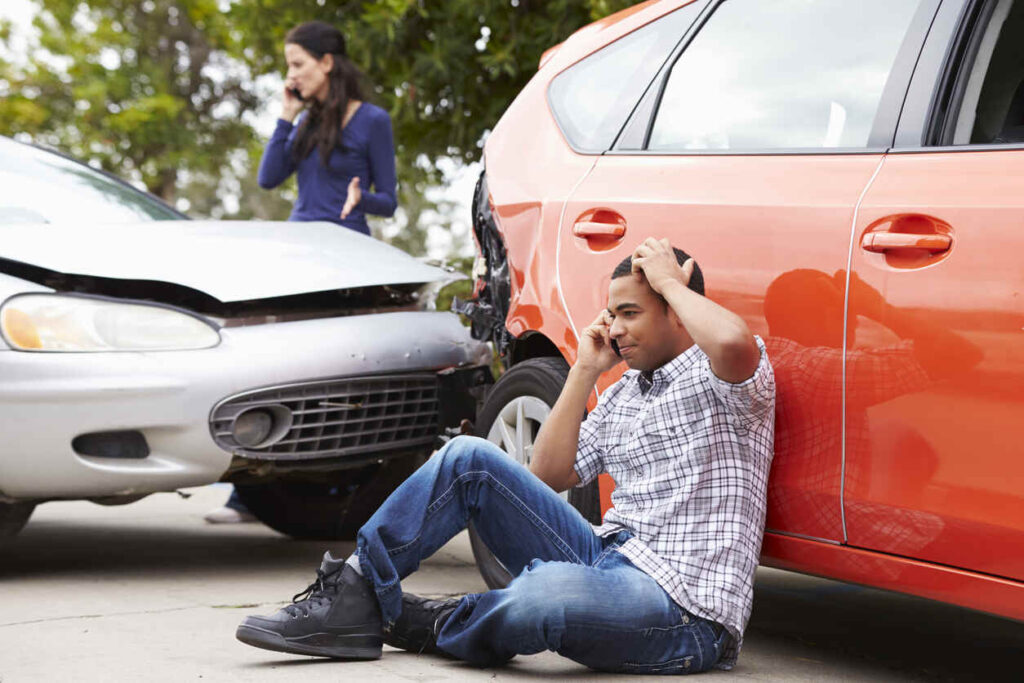 Person sitting on the ground after a rental car accident in Chicago, speaking on the phone with visible car damage in the background.